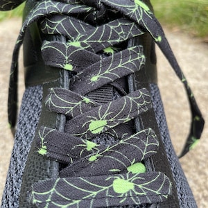 Może przedstawiać: Zbliżenie czarnego buta sportowego z czarnymi i zielonymi sznurowadłami w wzór pajęczyny. Sznurowadła są zawiązane w kokardę. But ma szarą cholewkę z siateczki i czarny język.