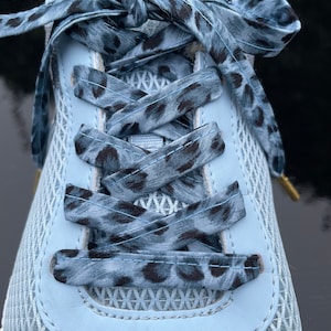 May include: Close-up of a light blue athletic shoe with matching leopard print laces. The shoe has a mesh upper and a white sole. The laces are tied in a bow.
