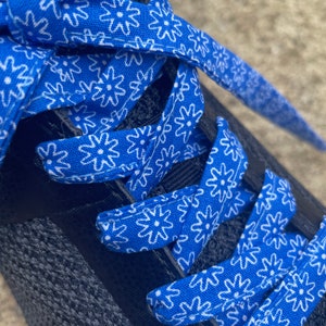 May include: Close-up of a black athletic shoe with bright blue and white floral patterned shoelaces. The laces are woven through the eyelets of the shoe, creating a striking contrast against the dark fabric of the shoe.
