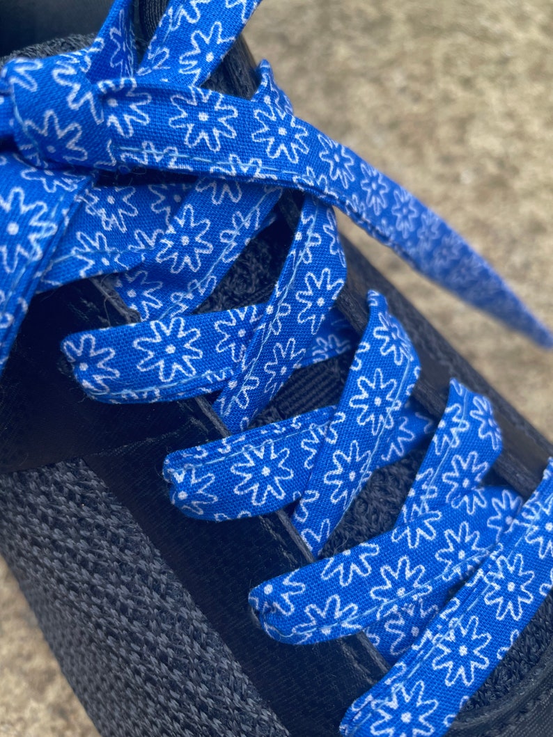 May include: Close-up of a black athletic shoe with bright blue and white floral patterned shoelaces. The laces are woven through the eyelets of the shoe, creating a striking contrast against the dark fabric of the shoe.