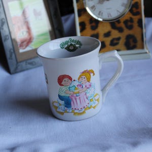 May include: A white ceramic mug featuring a Cabbage Patch Kids logo on the interior rim and a colorful illustration on the exterior showing two children sitting at a pink table sharing a large ice cream sundae with a spoon and blue straws.