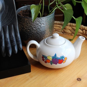 Pode incluir: Uma chaleira de cerâmica branca com um desenho de frutas, com tampa e bico. A chaleira está sobre uma superfície de madeira, ao lado de uma escultura de mão preta e uma planta em vaso. Capacidade de aproximadamente 1 litro.
