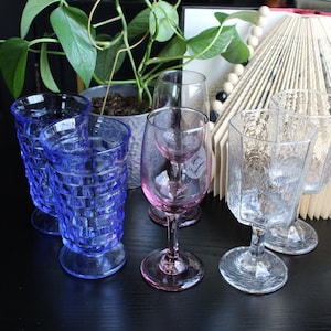 May include: A collection of glassware, featuring blue patterned tumblers, a pink wine glass, and clear textured glasses. A potted plant and a decorative book fan are in the background, all on a dark surface.