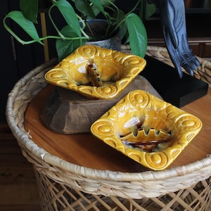 Puede incluir: Dos ceniceros cuadrados vintage con un esmalte amarillo mostaza. Cada cenicero tiene un borde decorativo en relieve y un reposacigarrillos incorporado. Los ceniceros se exhiben sobre una superficie de madera, encima de una mesa de mimbre tejida.