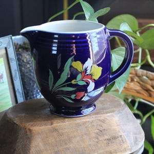 May include: A cobalt blue ceramic pitcher with a white interior and a handle. The pitcher is decorated with a floral design featuring yellow, white, and red flowers with green leaves. It sits on a wooden surface.