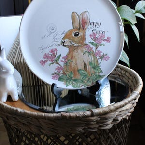 Könnte beinhalten: Weißer Keramik-Oster-Teller mit einem braunen Hasen, rosa Blumen und der Aufschrift "Happy Easter". Der Teller steht auf einem schwarzen Ständer, auf einem Tisch mit einem Weidenkorb. Durchmesser ca. 25 cm.