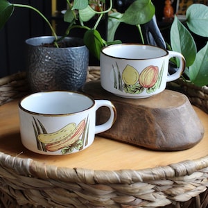 May include: Two white ceramic mugs with fruit illustrations. One mug features a lemon and a peach, the other a banana and a peach. Both mugs have a brown rim and handle. The mugs are on a wooden surface, with a woven basket base.