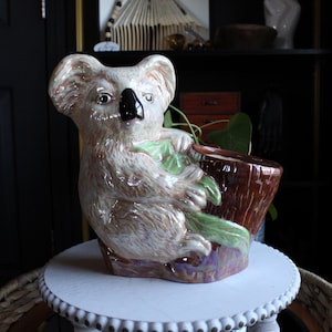 Puede incluir: Una maceta de cerámica con forma de oso koala con una maceta marrón con forma de tronco de árbol. El koala es de color marrón claro con nariz y ojos negros y hojas verdes. La maceta está sobre una mesa redonda blanca.