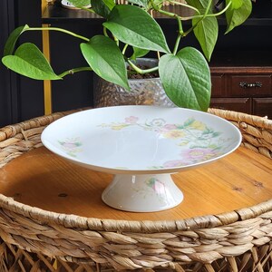 May include: White ceramic cake stand with a floral design in pink, yellow, and green. The stand has a wide, flat top and a short, pedestal base. The cake stand is displayed on a round, woven table.