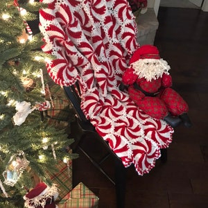 May include: A red and white crocheted blanket with a peppermint swirl design is draped over a black wooden chair. A small Santa Claus doll is sitting on the blanket.