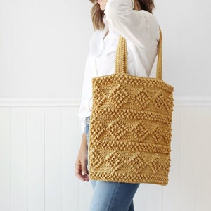 May include: A woman is holding a crocheted tote bag with a geometric pattern. The bag is a mustard yellow color and has a textured surface.