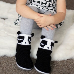 May include: A pair of black and white crocheted panda slippers with a white fur trim. The slippers are worn by a child sitting on a white fluffy rug.