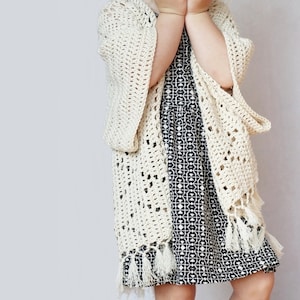 May include: A white crocheted cardigan with fringe trim. The cardigan is open in the front and has a loose fit. The cardigan is being worn over a black and white patterned dress.
