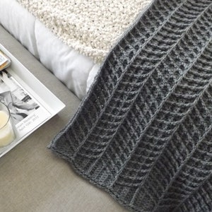 May include: A gray crocheted throw blanket with a chevron pattern. The blanket is folded and draped over a white couch.