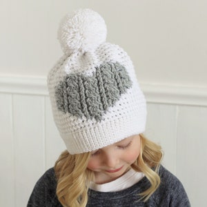 May include: White crochet beanie with a gray heart design and a white pom pom on top.