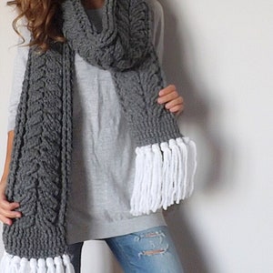 May include: A grey knitted scarf with a cable knit pattern and white fringe. The scarf is worn by a person wearing a grey sweater and blue jeans.