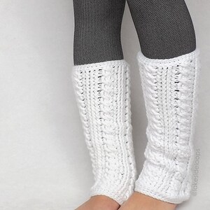 May include: White crocheted leg warmers with a cable knit pattern. The leg warmers are worn over black tights.