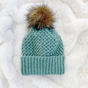 May include: A teal blue crocheted beanie with a brown faux fur pom pom. The beanie has a textured stitch pattern and a ribbed brim.