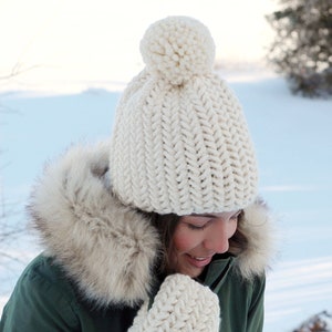 Pode incluir: Um chap&eacute;u de malha branco com um pompom no topo e luvas de malha brancas combinando. O chap&eacute;u e as luvas s&atilde;o usados por uma pessoa com uma jaqueta verde com capuz forrado de pele. A pessoa est&aacute; em p&eacute; em uma paisagem nevada.