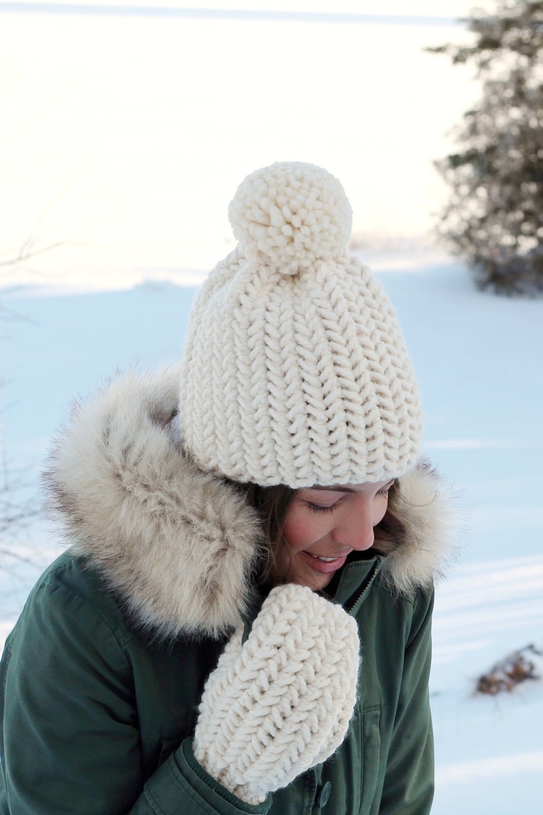 Pode incluir: Um chap&eacute;u de malha branco com um pompom no topo e luvas de malha brancas combinando. O chap&eacute;u e as luvas s&atilde;o usados por uma pessoa com uma jaqueta verde com capuz forrado de pele. A pessoa est&aacute; em p&eacute; em uma paisagem nevada.