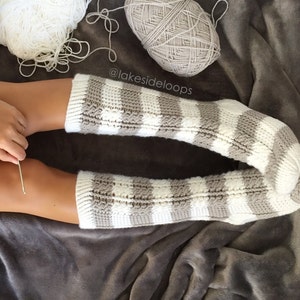 May include: A pair of cozy striped knit socks in white and gray with a cable knit pattern. The socks are on a gray fuzzy blanket. The text "@lakesideloops" is visible in the image.