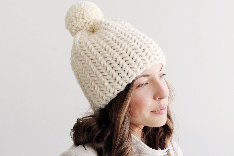 Pode incluir: Um gorro de malha branco com um pompom branco no topo. O gorro &eacute; feito com um fio grosso e tem uma textura canelada.