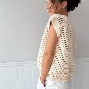 May include: Cream-colored, open-weave crochet top. The sleeveless garment has a loose fit and a textured appearance. The top is shown on a person wearing white pants. The background is a plain white wall.