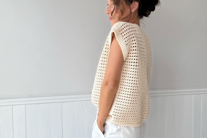 May include: Cream-colored, open-weave crochet top. The sleeveless garment has a loose fit and a textured appearance. The top is shown on a person wearing white pants. The background is a plain white wall.