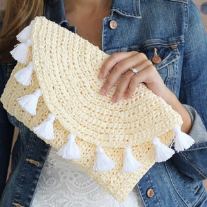 May include: A cream crocheted clutch bag with white tassel accents. The bag is made from a woven straw-like material and has a rounded shape.