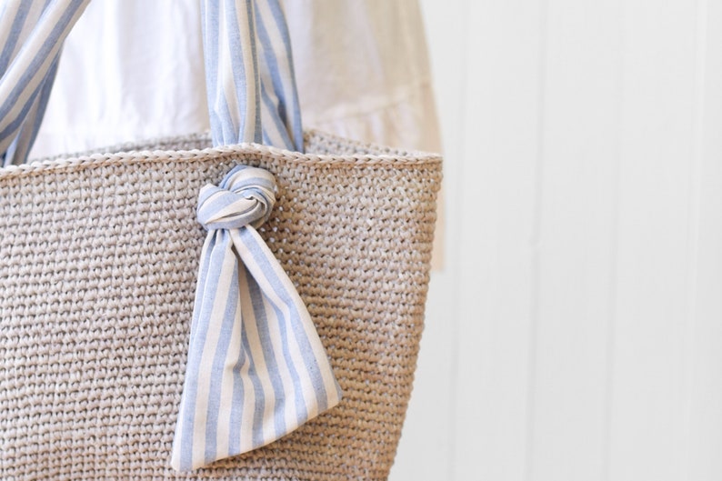 May include: A beige crocheted tote bag with blue and white striped fabric handles tied in a knot at the top.
