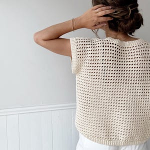 May include: Cream-colored, open-weave crochet top. The sleeveless top has a rounded neckline and a slightly curved hem. The texture of the crochet creates a grid-like pattern. The top is worn over a white garment.