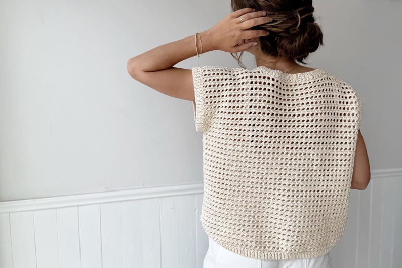 May include: Cream-colored, open-weave crochet top. The sleeveless top has a rounded neckline and a slightly curved hem. The texture of the crochet creates a grid-like pattern. The top is worn over a white garment.