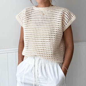May include: Cream-colored, open-weave crochet top with short sleeves. The top has a round neckline and a loose, boxy fit. Paired with white pants. The crochet design creates a textured, airy look, suitable for warm weather.