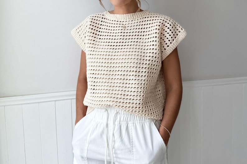 May include: Cream-colored, open-weave crochet top with short sleeves. The top has a round neckline and a loose, boxy fit. Paired with white pants. The crochet design creates a textured, airy look, suitable for warm weather.