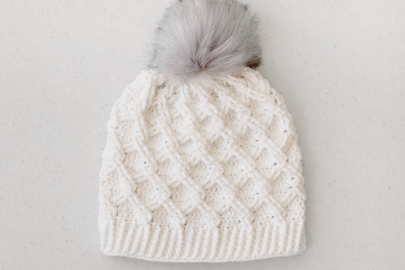 May include: A white crocheted beanie with a fluffy white pom pom on top. The beanie has a textured, cable knit design.