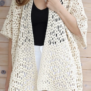 May include: A crocheted cream-colored cardigan with a loose fit and short sleeves. The cardigan is made with a lacy stitch pattern and has a slightly longer back than front.