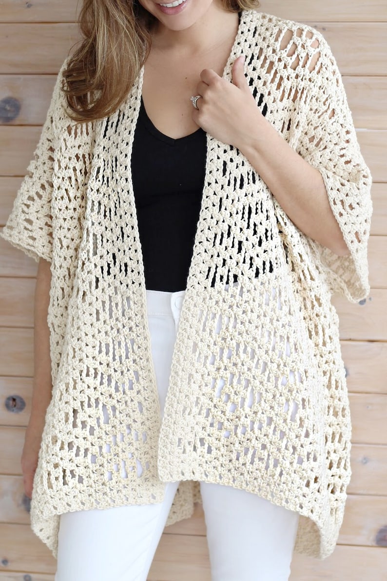 May include: A crocheted cream-colored cardigan with a loose fit and short sleeves. The cardigan is made with a lacy stitch pattern and has a slightly longer back than front.