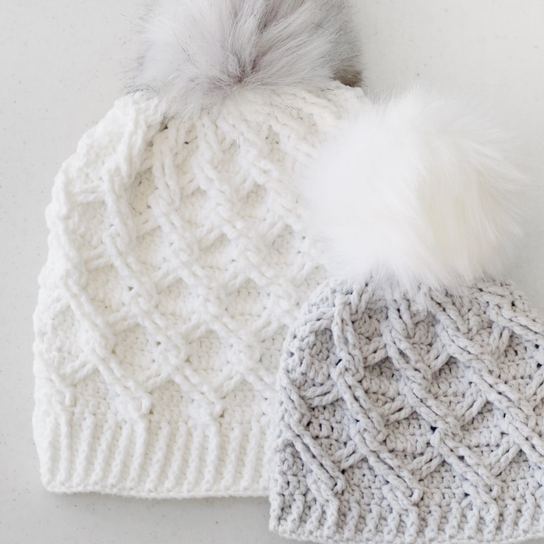 May include: Two crocheted hats with faux fur pom poms. The hat on the left is white with a diamond pattern. The hat on the right is gray with a cable knit pattern.
