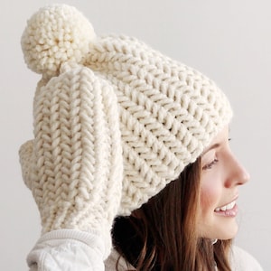 Pode incluir: Um chap&eacute;u branco de malha com um pompom no topo e luvas combinando. O chap&eacute;u e as luvas s&atilde;o feitos com um padr&atilde;o de ponto chevron.