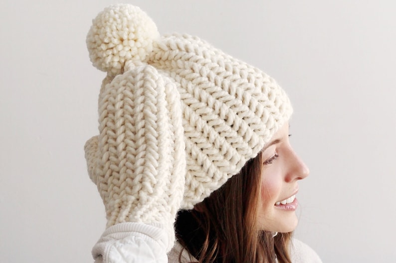 Pode incluir: Um chap&eacute;u branco de malha com um pompom no topo e luvas combinando. O chap&eacute;u e as luvas s&atilde;o feitos com um padr&atilde;o de ponto chevron.