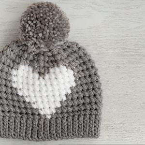 May include: Gray crocheted beanie with a white heart design and a fluffy gray pom pom on top.