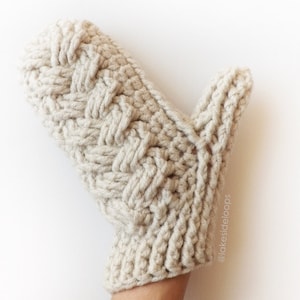 May include: A beige crocheted mitten with a cable knit pattern. The mitten is shown on a hand, with the thumb extended. The text "@lakesideloops" is visible on the mitten.