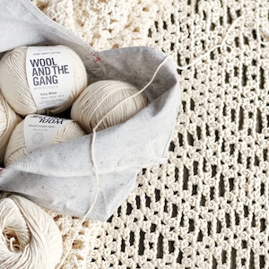 May include: Four balls of ivory white yarn in a grey canvas bag with the text "Wool and the Gang" on the label. The yarn is on a white crocheted surface.