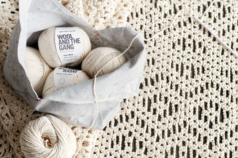 May include: Four balls of ivory white yarn in a grey canvas bag with the text "Wool and the Gang" on the label. The yarn is on a white crocheted surface.