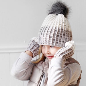 May include: A child wearing a white and brown crocheted hat with a gray pom pom and matching crocheted mittens.