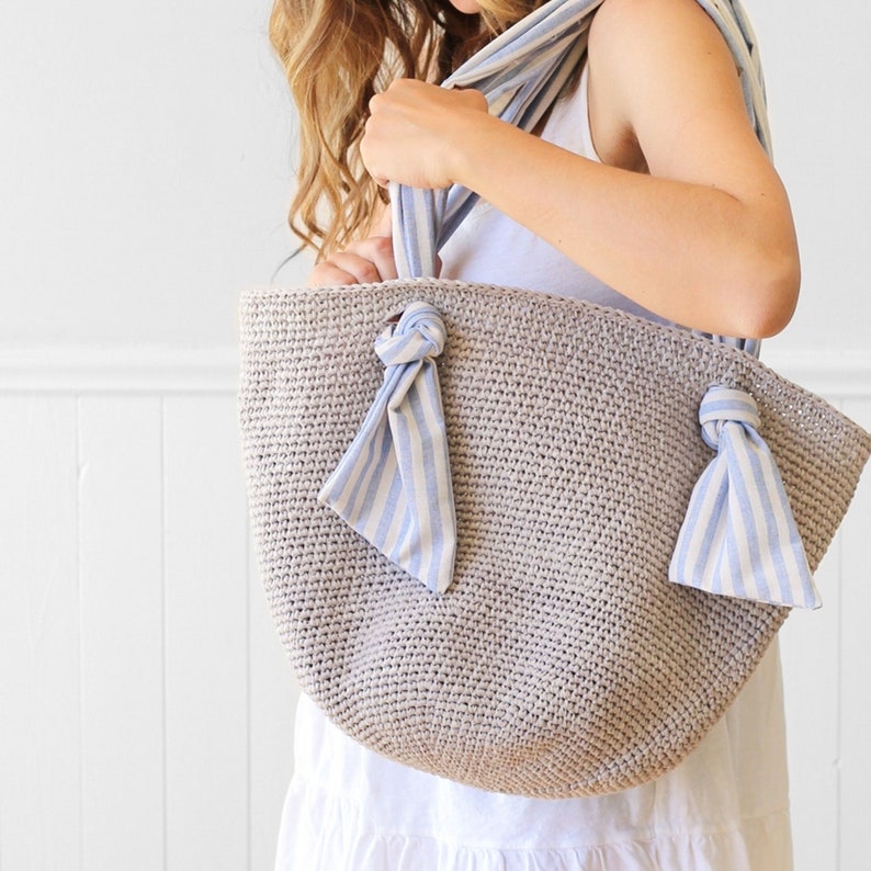 May include: A large, crocheted tote bag with a light gray color. The bag has two blue and white striped fabric bows attached to the sides.