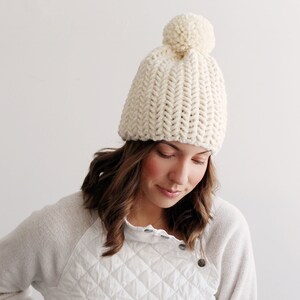 Pode incluir: Um gorro de malha branco com um pompom no topo. O gorro &eacute; feito com um fio grosso e tem uma textura canelada.