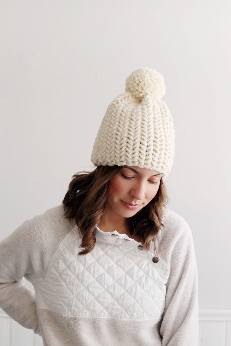 Pode incluir: Um gorro de malha branco com um pompom no topo. O gorro &eacute; feito com um fio grosso e tem uma textura canelada.