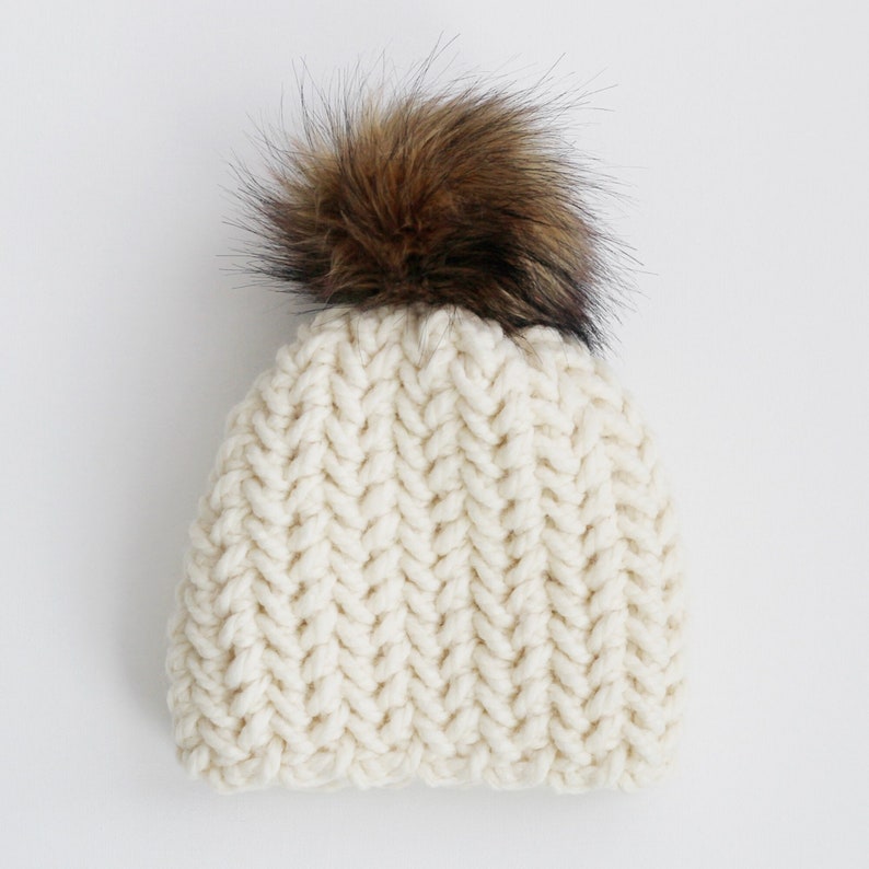 Pode incluir: Um gorro de malha branco com um pompom de pele sint&eacute;tica marrom.