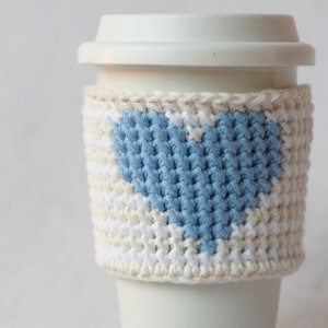 Puede incluir: Taza de café blanca con un acogedor de punto de crochet azul y blanco en forma de corazón.
